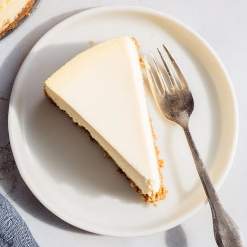 A chilled slice of New York Cheesecake on a dessert plate, topped with a fresh strawberry and sweet vanilla sauce, ready to eat.