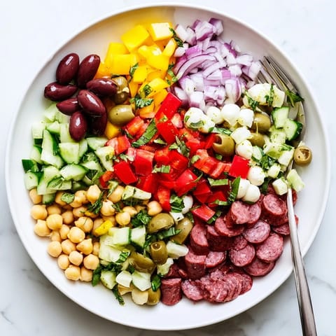 A close-up of Crunchy Antipasto Chopped Salad in a white bowl, featuring vibrant red tomatoes, green cucumber, cubes of salami, and mozzarella pearls tossed in a zesty Italian dressing.