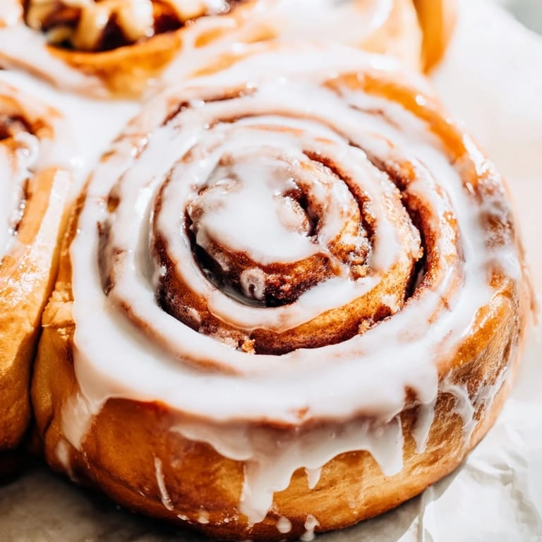 Steaming Cinnamon Rolls sit in a ceramic baking dish, their gooey brown sugar and cinnamon filling peeking through golden spirals under a thick white icing.