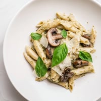 Golden-brown sautéed mushrooms tossed with penne in a vibrant, creamy basil pesto sauce, garnished with fresh basil and Parmesan.